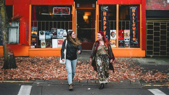 Two people walking past a red storefront with autumn leaves on the ground.
