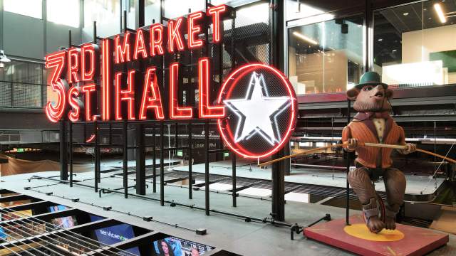 Neon sign reading “3rd Street Market Hall” glows brightly inside a modern indoor space, with a large white star outlined in red next to it. To the right, a whimsical statue of a dressed-up bear in a striped suit, monocle, and bowler hat balances on a unicycle while holding a tightrope pole.