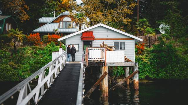 Two people stand on a narrow walkway leading to a small oceanfront house on stilts above the water, surrounded by trees and fall foliage.