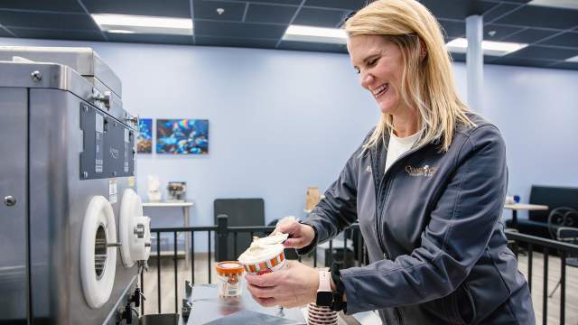 A blonde haired woman scopes ice cream from a manufacturing size ice cream machine into pint cups.