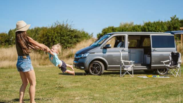 A woman swinging a toddler by his arms in front of a camper van.