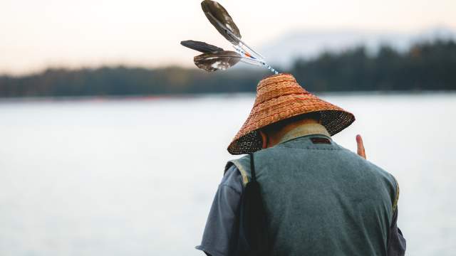 Person viewed from behind wearing a woven cedar hat decorated with feathers while standing near the shoreline.