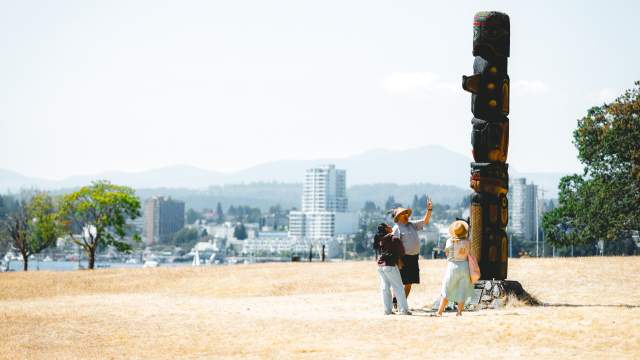 Three people stand at the base of a totem pole on Saysutshun with Nanaimo’s skyline visible across the harbour in the background.