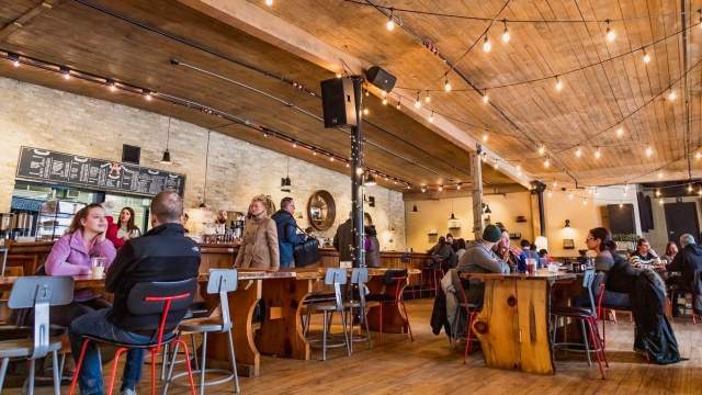 A wide interior view of a busy café or taproom with a high wooden ceiling and string lights. Groups of people sit and stand around wooden tables and benches, talking and socializing. The space feels warm and lively, with a bar counter visible in the background and natural wood features throughout.