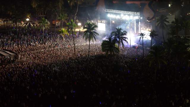 Aerial photo of crowd watching an outdoor concert at night. The stage is brightly lit which illuminates parts of the audience and palm trees