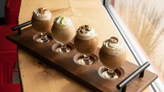 A wooden flight board holding four small glass cups of iced coffee or dessert drinks topped with whipped cream and garnishes, placed on a wooden counter by a window.
