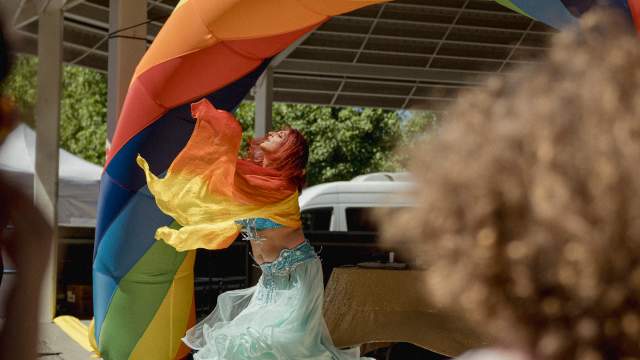 Performer in flowing costume dancing beneath a rainbow arch on a stage at a Pride event, with audience in the foreground.