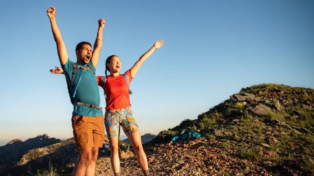 Man and Woman wearing hiking gear with their arms up and looking excited to reach the top of a mountain