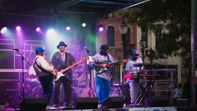 A blues band performing on an outdoor stage at Michigan BluesFest, with guitars, drums, and a cheering crowd.