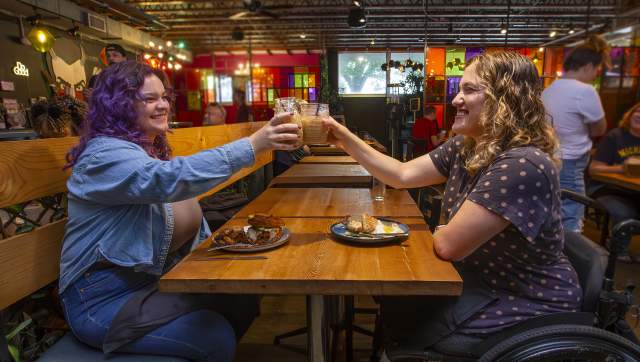 A woman with purple hair clinks glasses with a smiling woman in a wheelchair above plates full of food at the People's Kitchen.