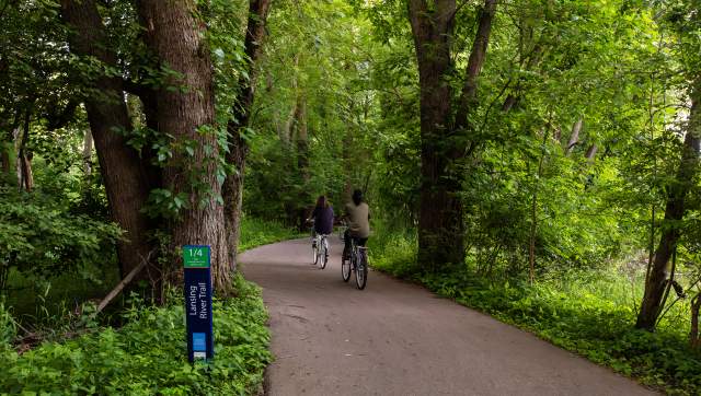 Two bikers riding on the Lansing River Trail surrounded by lush green plants