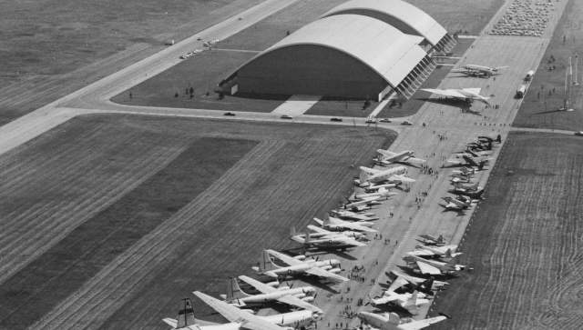 An exterior view of the National Museum of the U.S. Air Force, as it appeared in the early 1970s, with numerous aircraft lined up on a runway adjacent to the museum building.