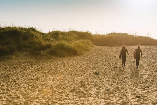 Two ladies walking across the beach with surfboards