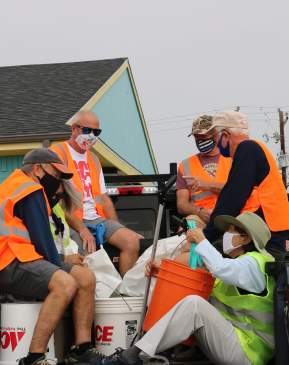 A group of five volunteers in orange safety vests sit in the bed of a truck with trash bags and buckets