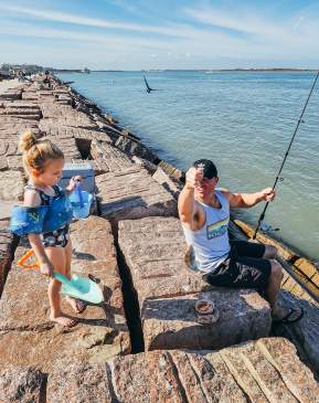 A father sitting on a jetty helps his young daughter learn how to fish.