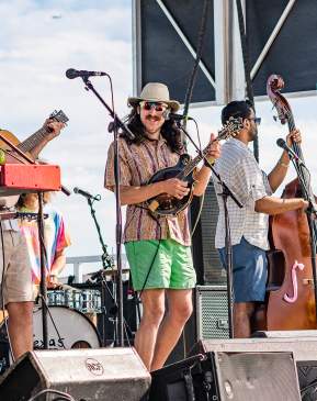 A four-person band performs on a stage. The one in the middle plays a small guitar-like instrument and smiles at the camera.