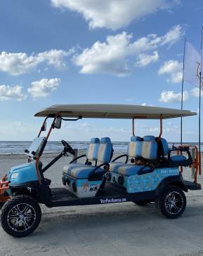 A light blue and patterned golf cart with two large flags sits on the beach.
