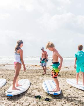 A group of five kids stand on surf boards resting on the sand while an instructor stands in front of them