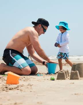 A man in a swimsuit kneels on the sand next to a child wearing sun gear and a hat. They pack sand in a blue bucket and build a sandcastle.