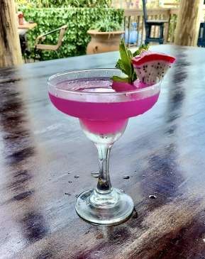 Fuschia-colored margarita with a dragonfruit wedge on the rim sits on a wooden table.