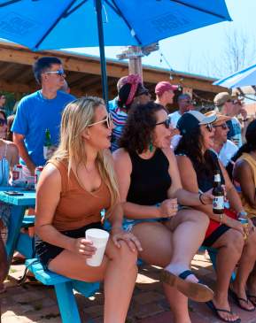 A diverse group of people sits at a picnic table with excited expressions as they watch an off-camera event
