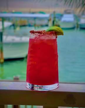 Glass of a red margarita with a lime on a ledge overlooking water.