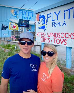 A couple in sunglasses pose together in front of a store called "Fly It! Port A"