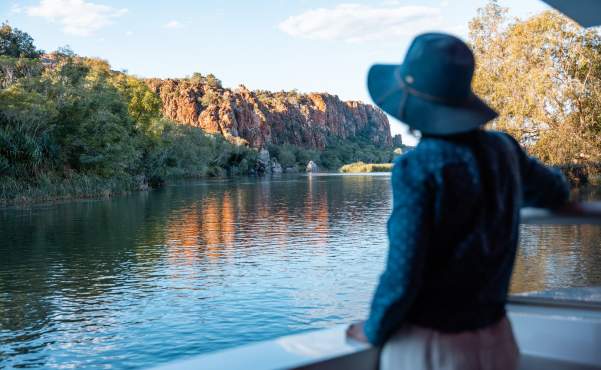 A woman enjoying the scenery on an Ord River cruise - the woman is standing on the vessel looking across the water at a cliff faced bathed in sunlight.