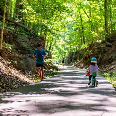 a child rides a bike on a paved trail beside a man running