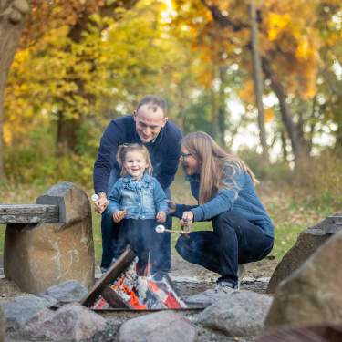 A white man, white woman and young white girl smile while roasting marshmallows over the fire surrounded by fall foliage at Picnic Point
