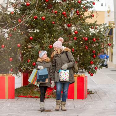 A mom and her daughter stand in front of a Christmas tree with bags of gifts