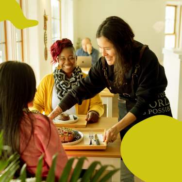 A woman sets a tray of food down on a table between two women