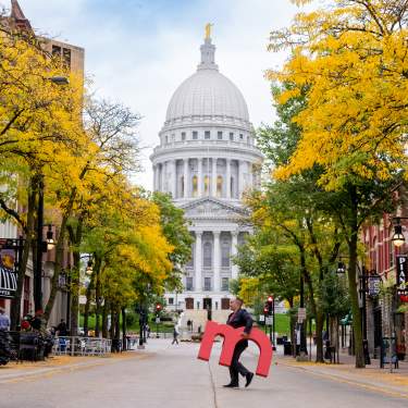 An image of a man walking across State Street in downtown Madison holding a large red "m" with the Capitol building in the background.