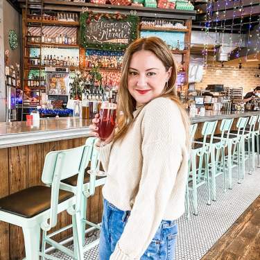 A white woman stands holding a holiday cocktail in front of the bar at Lucille, which has been transformed into a holiday wonderland