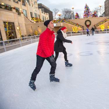 A man and woman ice skate at the Edgewater with Christmas trees and decor in the background.