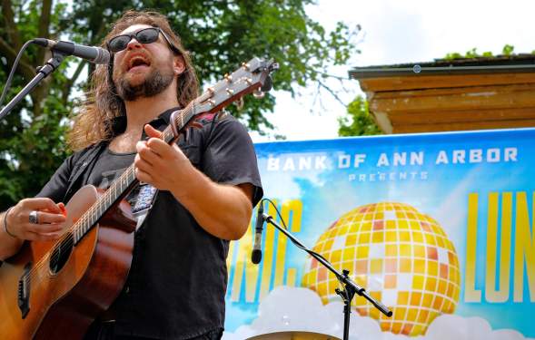 Man wears sunglasses and sings into a microphone while playing acoustic guitar. He stands in front of a blue banner with a yellow disco ball printed on the banner.