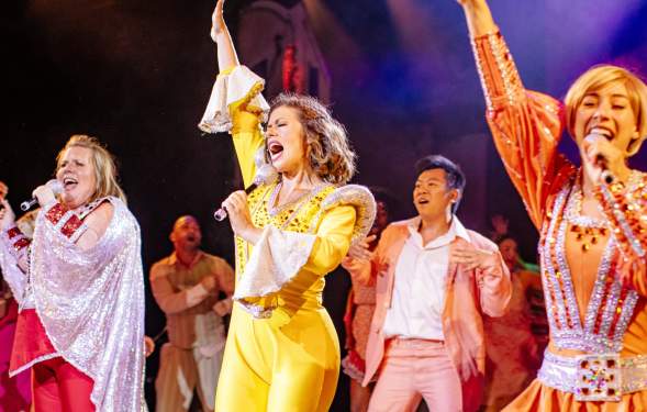 Three women dressed up in 70s outfits sing on stage with their hands raised in unison. Behind the three women is a man dancing backup. The group is singing a song.