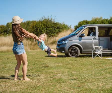 A woman swinging a toddler by his arms in front of a camper van.