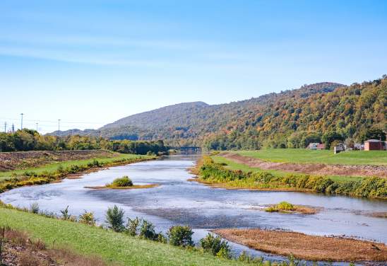 River winding through a grassy valley with tree-covered mountains in the background under a clear blue sky.