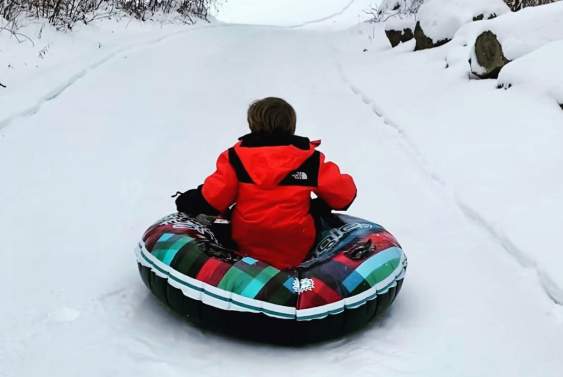 A boy in a red coat sits in a colorful, inflated snow tube and slides down a snow-covered hill.
