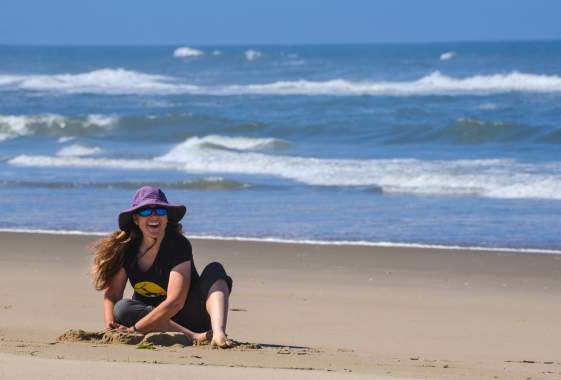 Woman sits building a sand castle on the beach with the ocean behind her. She is smiling under a large, purple brimmed hat.