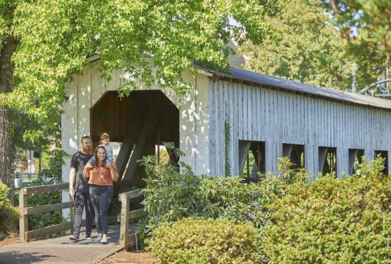 A young family of three exit the white, wooden Centennial Covered Bridge with trees overhead.