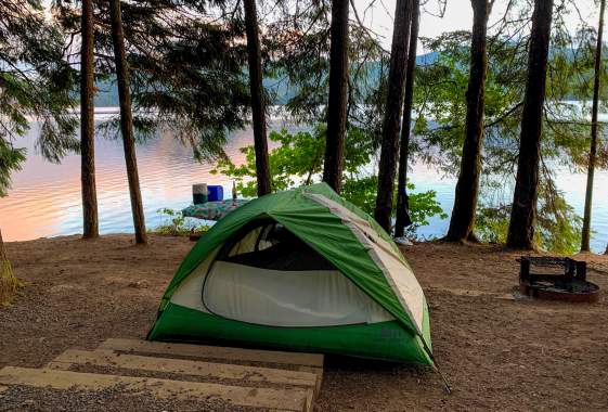 A green and white tent sits beside a lake with tall trees and sunrise colors reflecting off the water.