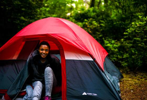 A woman sits smiling at the entrance of a small red tent in the forest.