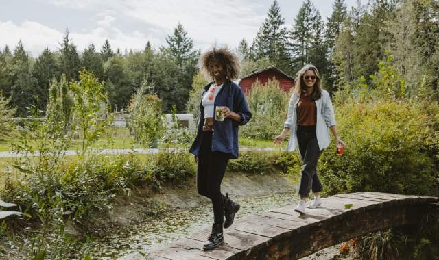 Two women walk across a small bridge, carrying glasses of cider.