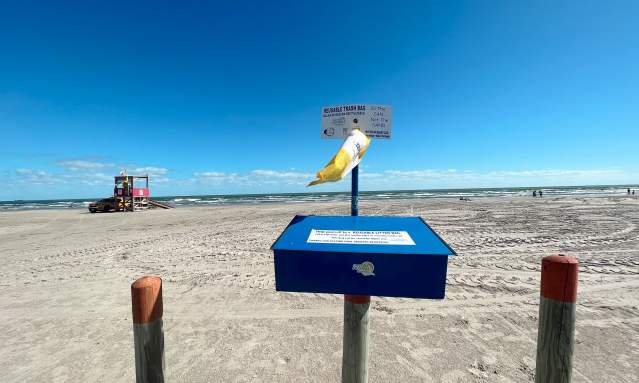 In the foreground is a blue metal box mounted on a wooden pole. From it hangs a yellow mesh bag. In the background is the beach and a lifeguard station.