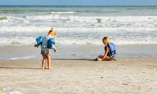 Two blonde toddlers, one in water wings and one in a life jacket, play on the beach by the water. one is kneeling in the sand, one is standing, walking towards the water