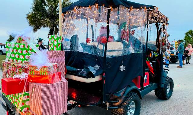 A golf cart is decked out with Christmas lights. On a trailer, it pulls a bundle of Christmas gifts.