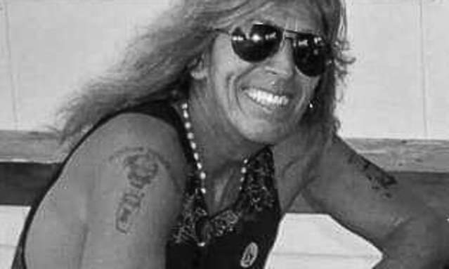 Black and white photo of a long-haired man in sunglasses leaning on a Conga drum.