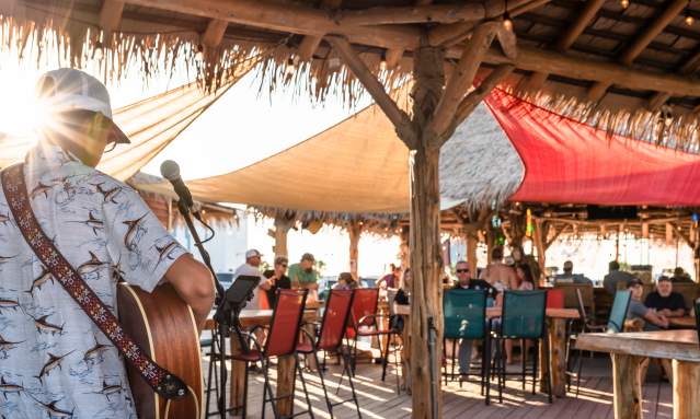 Photo from behind a musician playing guitar towards a tiki bar